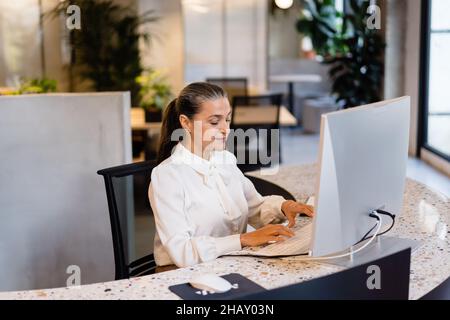 Female manager in elegant wear typing on computer while sitting at desktop during work in modern light office Stock Photo