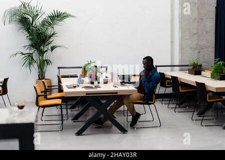 Full body side view of focused African American male manager typing on netbook while sitting at desktop in spacious workspace Stock Photo