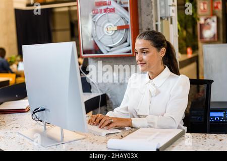 Smiling female manager in elegant wear typing on computer while sitting at desktop during work in modern light office Stock Photo