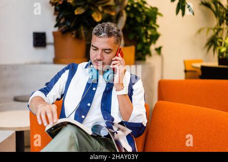 Focused male worker with headphones reading magazine and talking on smartphone while sitting on armchair in modern office during work Stock Photo
