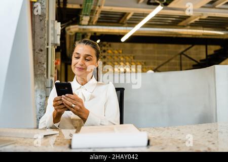 Smiling female manager in elegant outfit browsing on mobile phone at desktop during work in modern light office Stock Photo