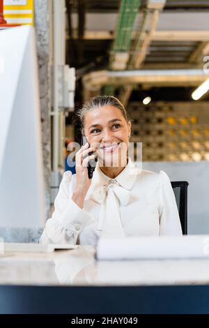 Smiling female manager in elegant outfit looking away while having phone call at desktop during work in modern light office Stock Photo