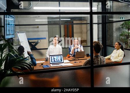 Through window group of colleagues sitting at table with laptops while working on business project during online meeting in boardroom Stock Photo