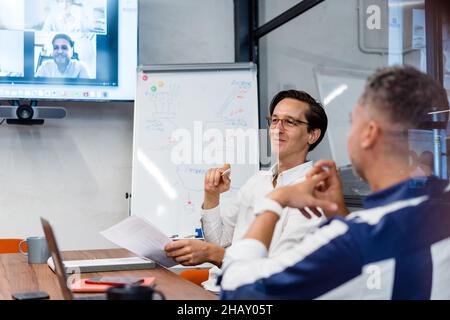 Coworkers sitting at table with gadgets and documents in modern workspace with white flip chat during online conference in office Stock Photo