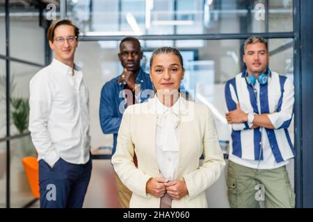 Female worker in formal wear looking at camera while standing near group of multiracial male coworkers in light modern office Stock Photo