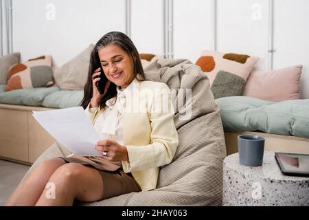 Positive female worker talking on cellphone and reading documents while sitting on bean bag during work in modern light office Stock Photo