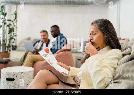 Woman drinking coffee and reading documents with charts while sitting on sofa near multiethnic male colleagues during work in modern office Stock Photo