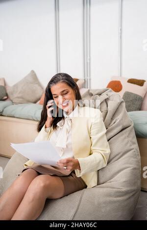 Positive female worker talking on cellphone and reading documents while sitting on bean bag during work in modern light office Stock Photo