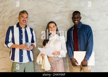 Group of positive multiracial coworkers with documents and laptop in hands looking at camera while standing together near wall Stock Photo