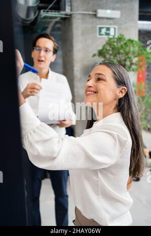 Side view of happy female worker taking notes on stickers attached on glass wall of room with colleagues while working in modern office Stock Photo