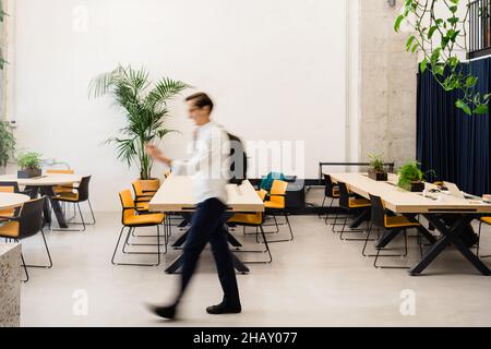 Full body side view of blurred male worker with backpack strolling in light modern workspace with tables during work in spacious workspace Stock Photo