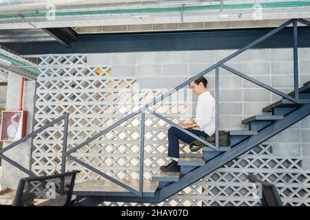 Full body side view of focused male worker with netbook sitting on staircase while working in light modern workspace during work Stock Photo