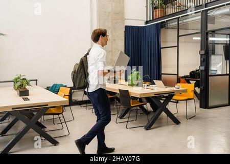 Side view of male worker with netbook in hand walking in work in light modern office with tables Stock Photo