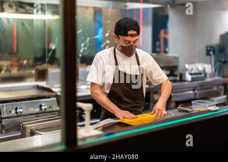 Side view of male cook in protective mask standing at stainless counter with steel tray of baked flatbread in restaurant open kitchen during pandemic Stock Photo