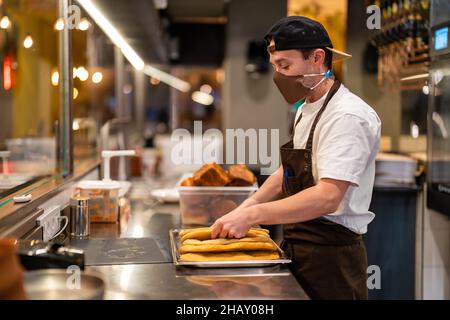 Side view of male cook in protective mask standing at stainless counter with steel tray of baked flatbread in restaurant open kitchen during pandemic Stock Photo