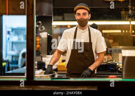Through glass of young male cook in apron cap and gloves protecting face with mask cooking in kitchen of restaurant and looking at camera Stock Photo