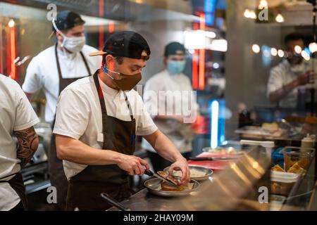 Through glass of focused male cook in uniform and protective mask adding ingredient to burger in kitchen with colleagues during coronavirus pandemic Stock Photo