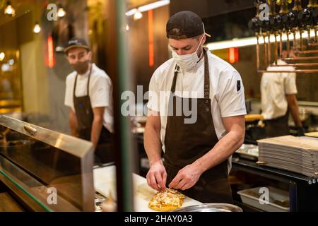 Male cook wearing uniform and mask for coronavirus prevention adding cheese on baked flatbread while working in restaurant Stock Photo