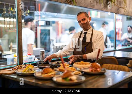 Stylish male waiter in uniform placing plates with assorted meals on table in restaurant with open kitchen during COVID pandemic Stock Photo