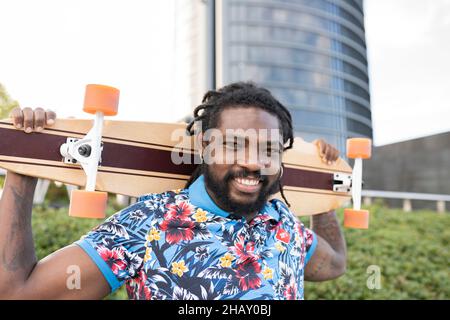 Delighted African American male in casual clothes with longboard behind head looking at camera while standing on street of city Stock Photo