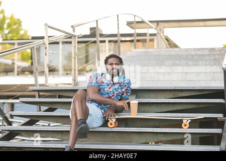 Thoughtful African American male in casual clothes with wireless headphones on neck looking into distance while sitting on stairs near building with t Stock Photo
