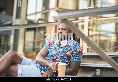 Thoughtful African American male in casual clothes with wireless headphones on neck looking into distance while sitting on stairs near building with t Stock Photo