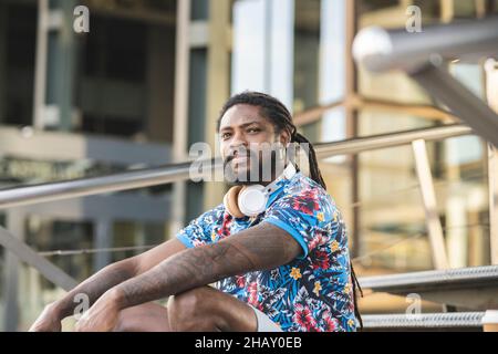 Thoughtful African American male in casual clothes with wireless headphones on neck looking into distance while sitting on stairs near building Stock Photo