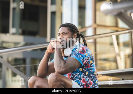 Thoughtful African American male in casual clothes with wireless headphones on neck looking into distance while sitting on stairs near building Stock Photo
