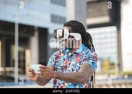 Side view of smiling trendy ethnic male with beard and cellphone listening to song from earphones on street Stock Photo