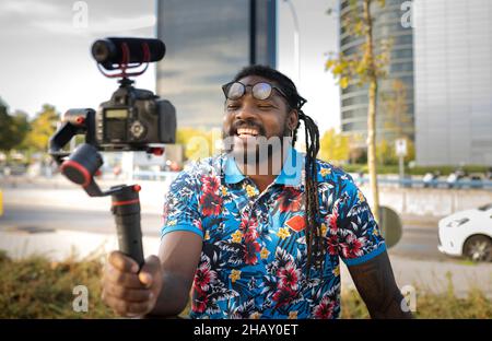 African American blogger with dreadlocks in eyeglasses shooting video on professional camera on street with trees in city Stock Photo