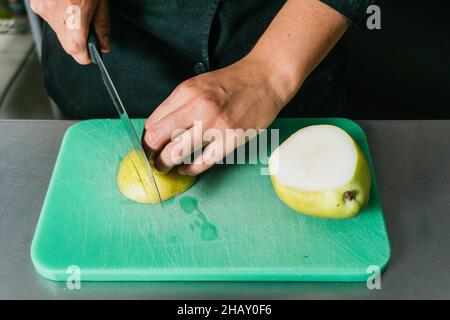Crop anonymous chef slicing fresh ripe pear on plastic cutting board with sharp knife in kitchen in restaurant Stock Photo