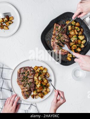Top view of crop anonymous people eating delicious beef chop with potatoes served on frying pan and plate on white background Stock Photo