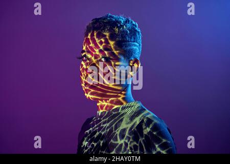 Charming African American female with short hair and in shadow projector light on face looking at camera on purple background in studio Stock Photo