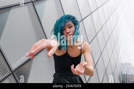 Charming female with blue hair looking at camera while performing dance near wall of contemporary building in daytime Stock Photo