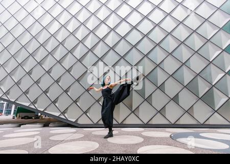 Full body of flexible Woman with blue hair in black clothes standing with raised leg and arms while dancing near modern building on street in daytime Stock Photo