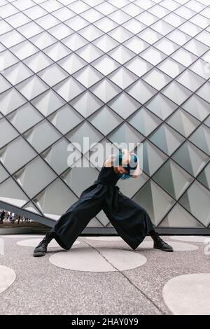 Young Woman with blue hair in black outfit performing contemporary dance near modern building on city street Stock Photo