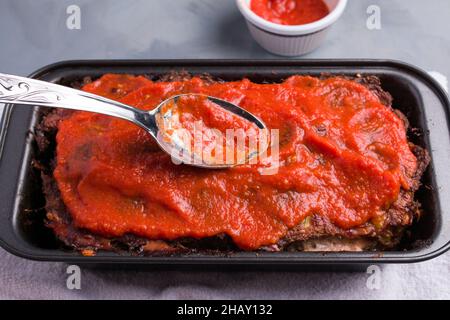 From above of spoon with delicious tomato sauce covering meat in black baking pan placed on gray table Stock Photo