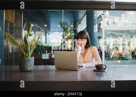 Young positive female in casual outfit sitting at table in cafe with cup of coffee watching at screen of laptop while having video call Stock Photo