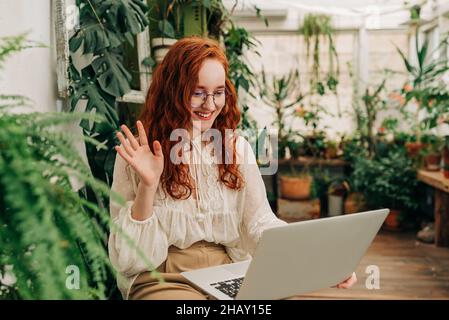 Positive red haired female remote worker in eyeglasses sitting among potted houseplants and waving to camera of netbook during video meeting Stock Photo