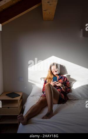 Blond lady sitting on bed and browsing smartphone while resting in attic Stock Photo