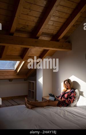 Side view of blond lady sitting on bed and browsing on tablet while resting in attic Stock Photo