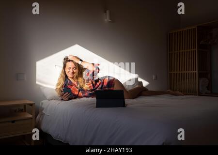 Laughing female using mobile phone in hand while lying on bed near tablet in light from window in dark bedroom Stock Photo