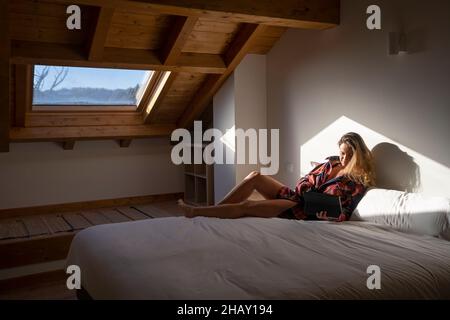 Side view of blond lady sitting on bed and browsing on tablet while resting in attic Stock Photo