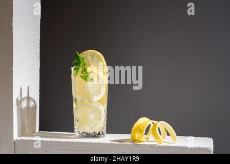 Glass of cold fresh lemonade with slices of lemon and leaves of mint placed on concrete block near lemon peel against grey background Stock Photo