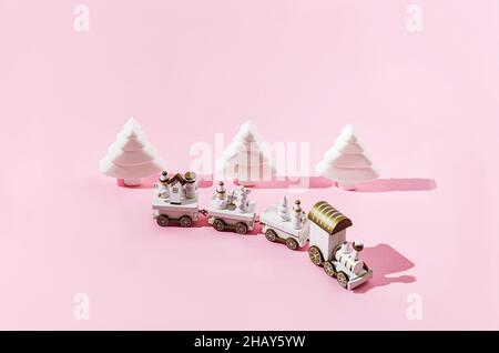 A white and golden Christmas train in the foreground, three white geometrical Christmas trees on pastel pink background. Minimal romantic creative con Stock Photo