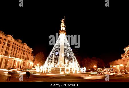 Tbilisi, Georgia - 12th december, 2021: Cars pass on roundabout leaving light trails with decoration lights on statue in freedom square while tourist Stock Photo