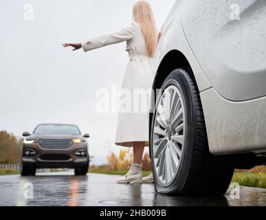 Back view of girl standing behind white vehicle with rear flat tire parked on edge of roadway and trying to stop oncoming SUV. Female driver of auto with damage tire giving stop sign to oncoming car. Stock Photo