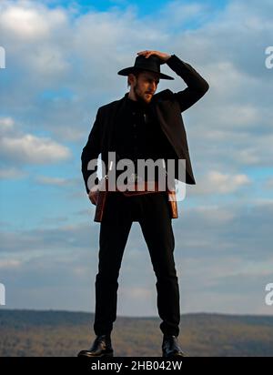 Sheriff or cowboy in black suit and cowboy hat. Man with west vintage pistol revolver gun and marshal ammunition. American western, sheriff. Stock Photo