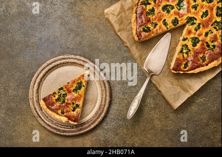 Piece of homemade quiche on ceramic rustic plate. Nearby is large pie on parchment paper with spatula. Top view Stock Photo