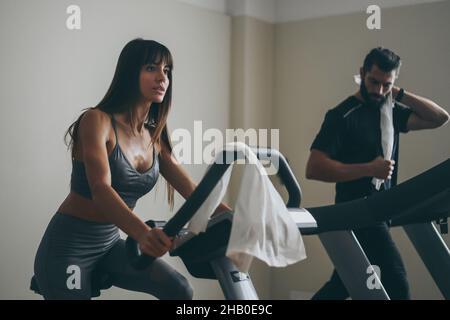 Determined young couple working out in gym. Fit boy and girl doing exercise with cyclette and tapis roulant. Beautiful sporty woman. Strong boys works Stock Photo